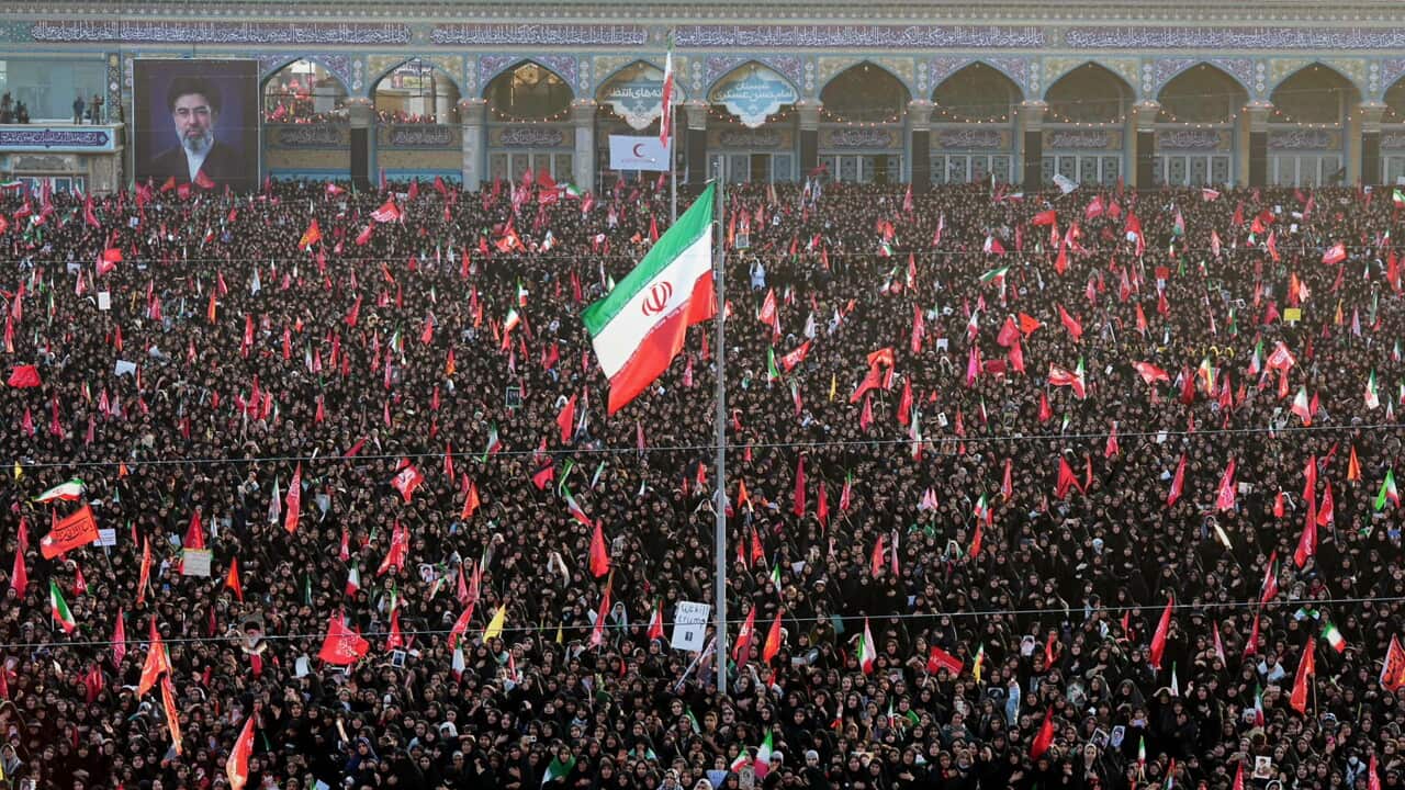 A large crowd surrounds a lone flagpole, flying an Iranian flag. People in the crowd carry red flags.