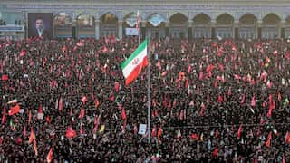 A large crowd surrounds a lone flagpole, flying an Iranian flag. People in the crowd carry red flags.