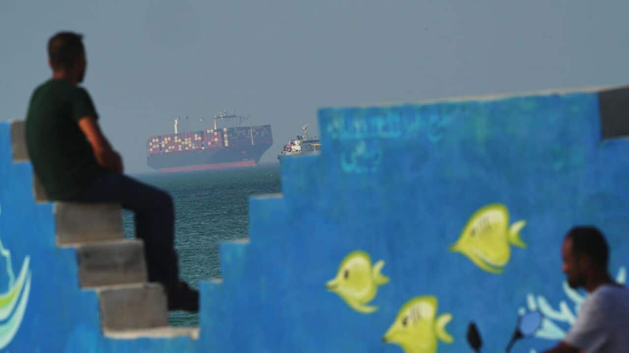 A man behind a mural depicting fishes at the shoreline watches as a cargo boat navigates the sea.
