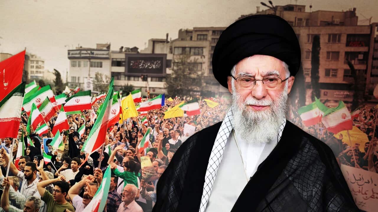 A cleric in a black robe and turban, with a grey beard and glasses on the right. With people protesting in the streets, holding up green, red and white Iran flags in the background on the left.