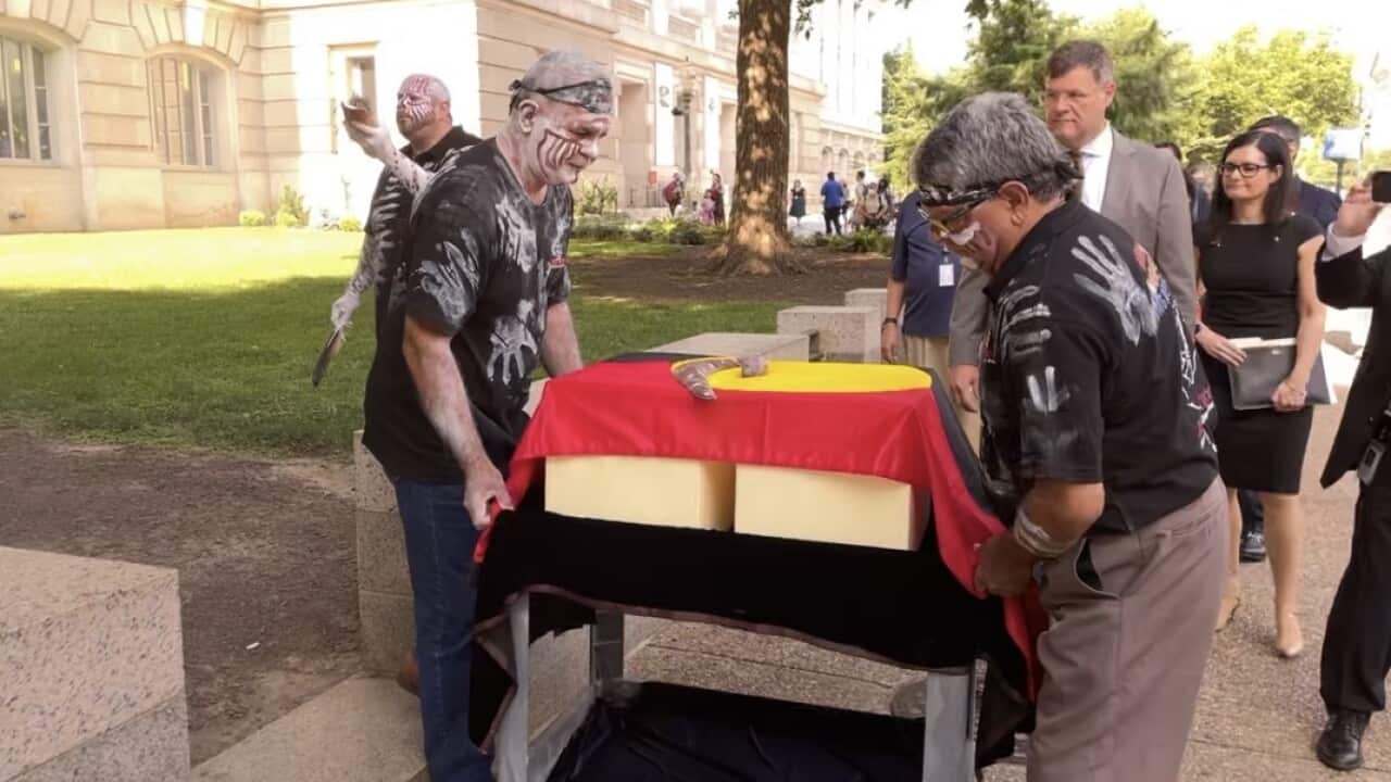 Two boxes draped in an Aboriginal flag with a boomerang on top. Several people surround the boxes, including several man in traditional ochre paint