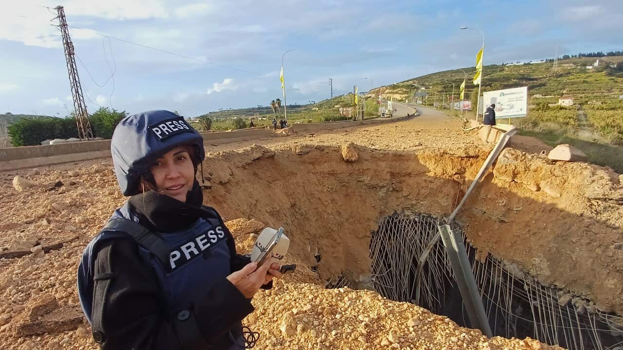 A woman wearing a press vest and helmet reports from a destroyed site