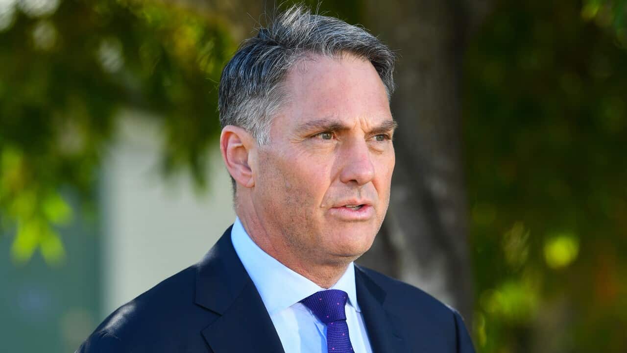 A man standing outside, wearing a blue suit and tie and white shirt standing partially side-on. The background is blurred.