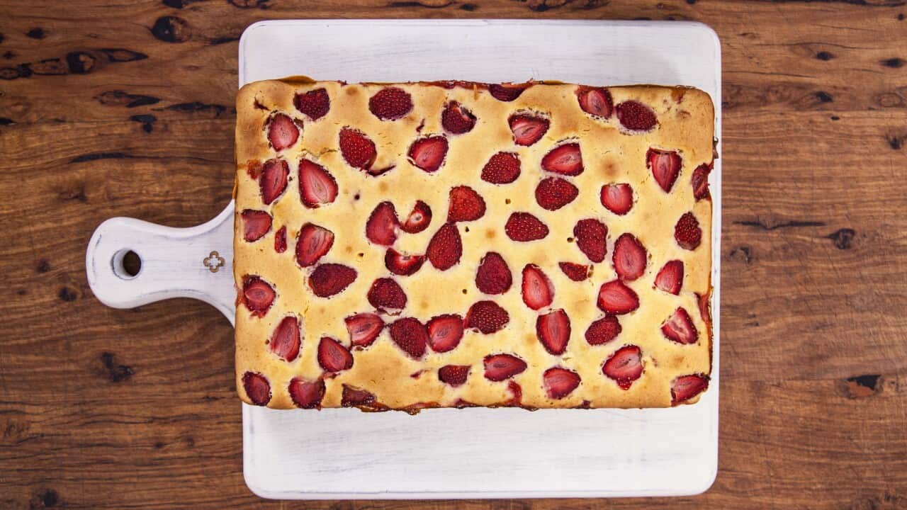 Seen from overhead, a rectangular cake studded with strawberries sits on a white paddle-shaped board.