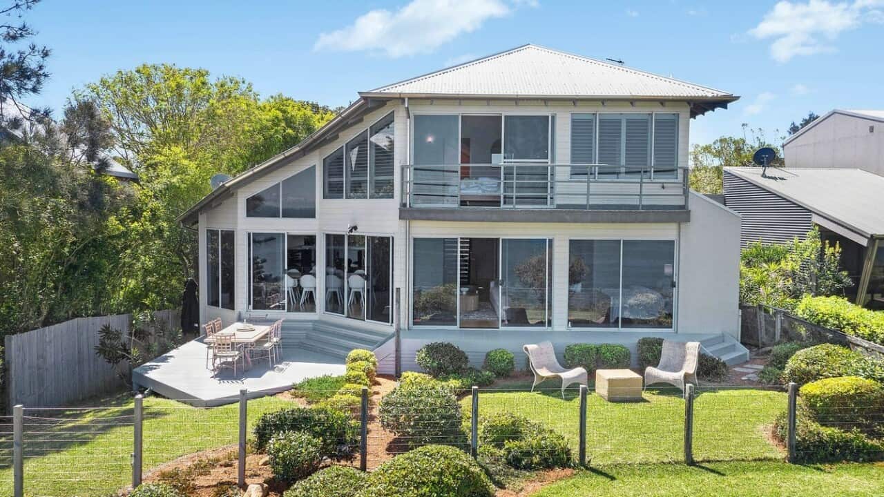 A double storey white weatherboard home, fenced off with a low clear fence.