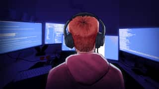 An illustration from behind a person with red hair wearing headphones, looking at multiple computer monitors displaying glowing blue screens filled with code in a dark room.