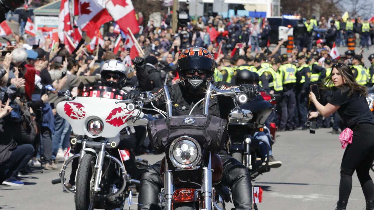 People riding motorcycles with a crowd standing behind them.