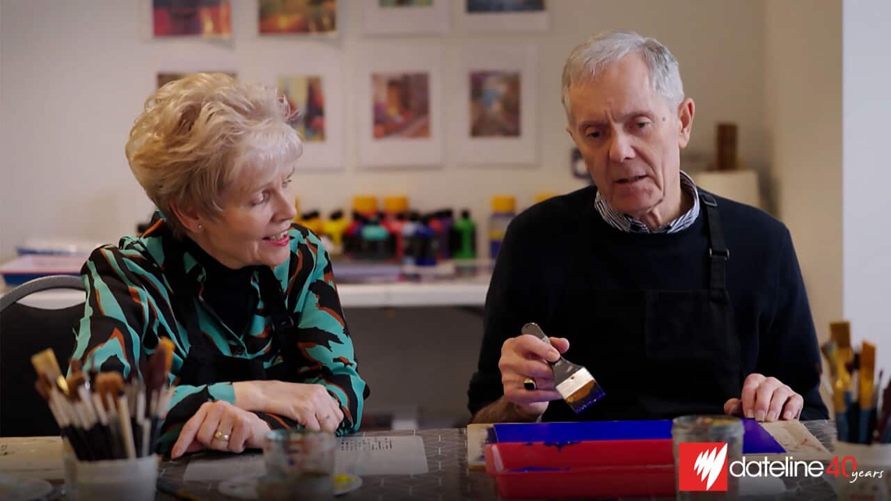 An elderly woman and a man sitting next to each other at a table in an art studio. The man is holding a painting brush and has a canvas lying in front of him
