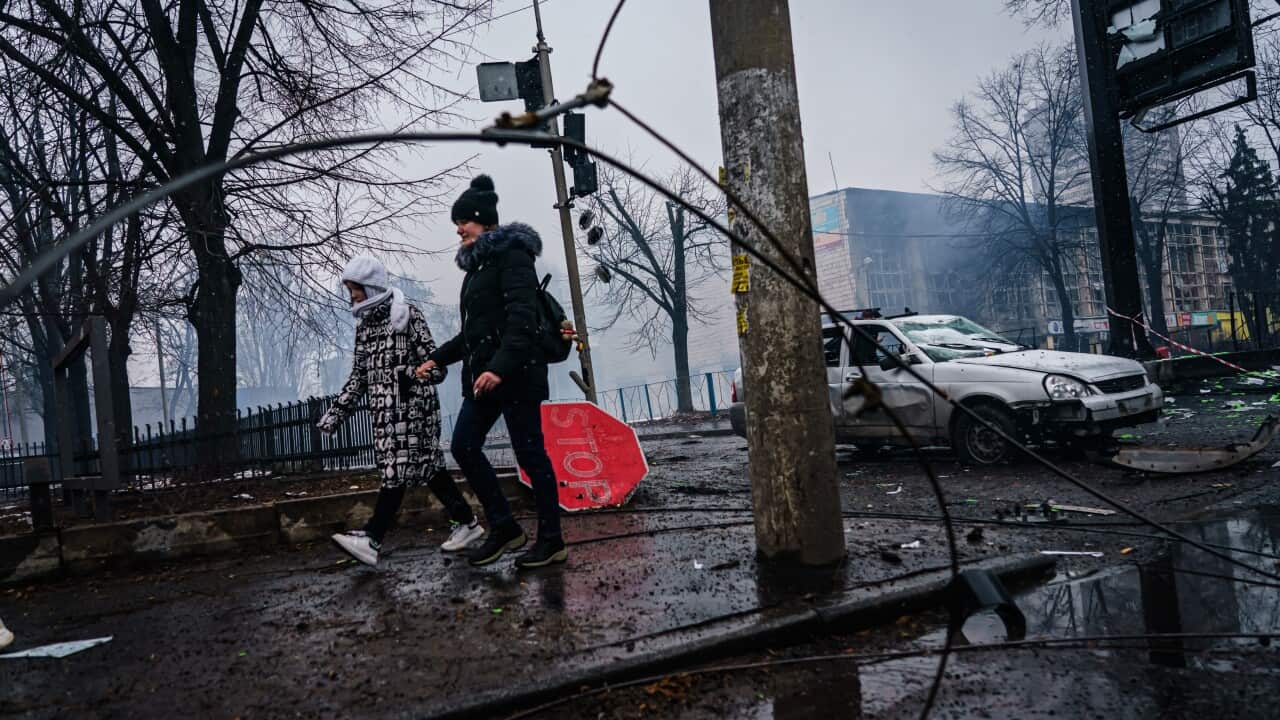 Two people are seen walking along a street with a damaged car and building in the background.