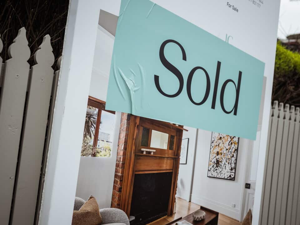 A sign for a recently sold house sits against a beige picket fence. The sign bears a picture of a red brick and wood fireplace.