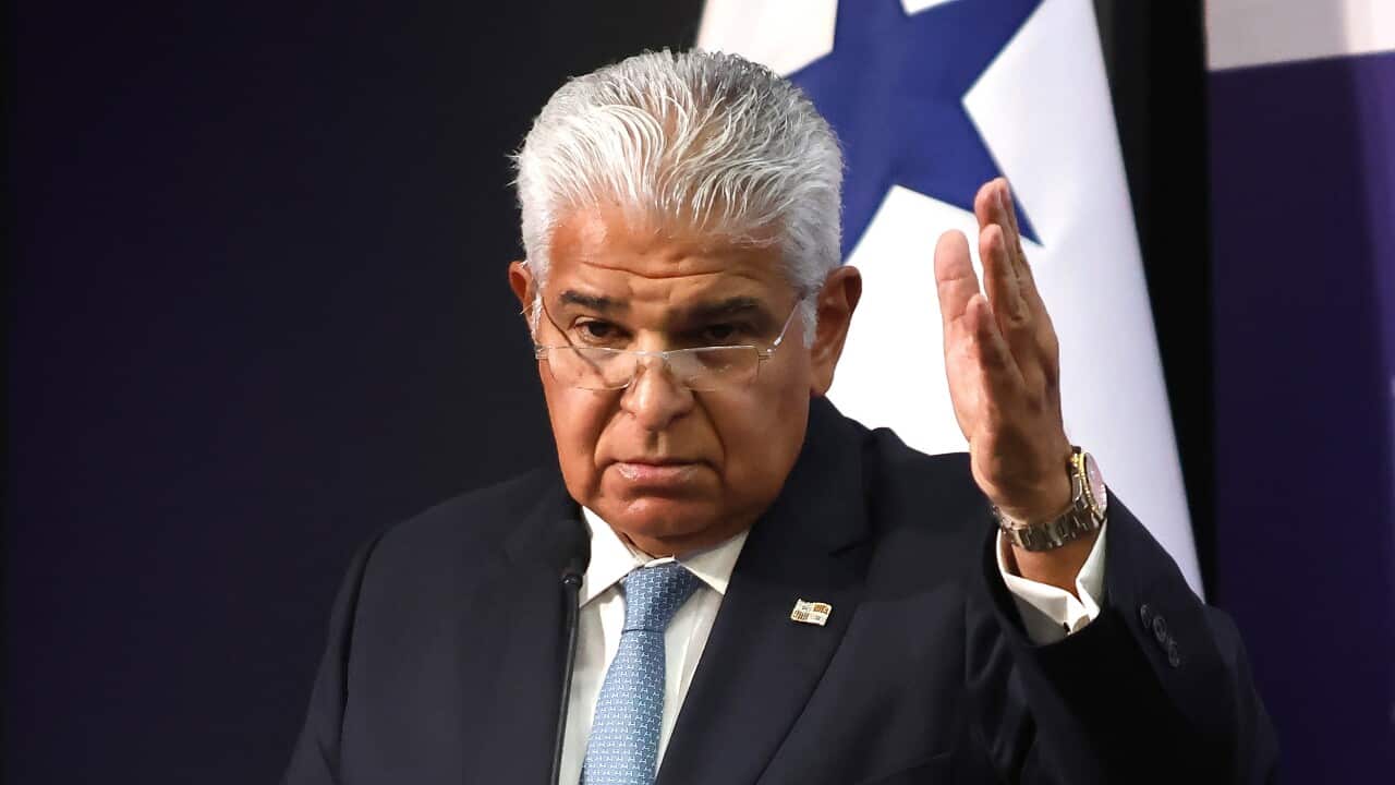 An elderly man in a dark suit speaks with one hand raised.