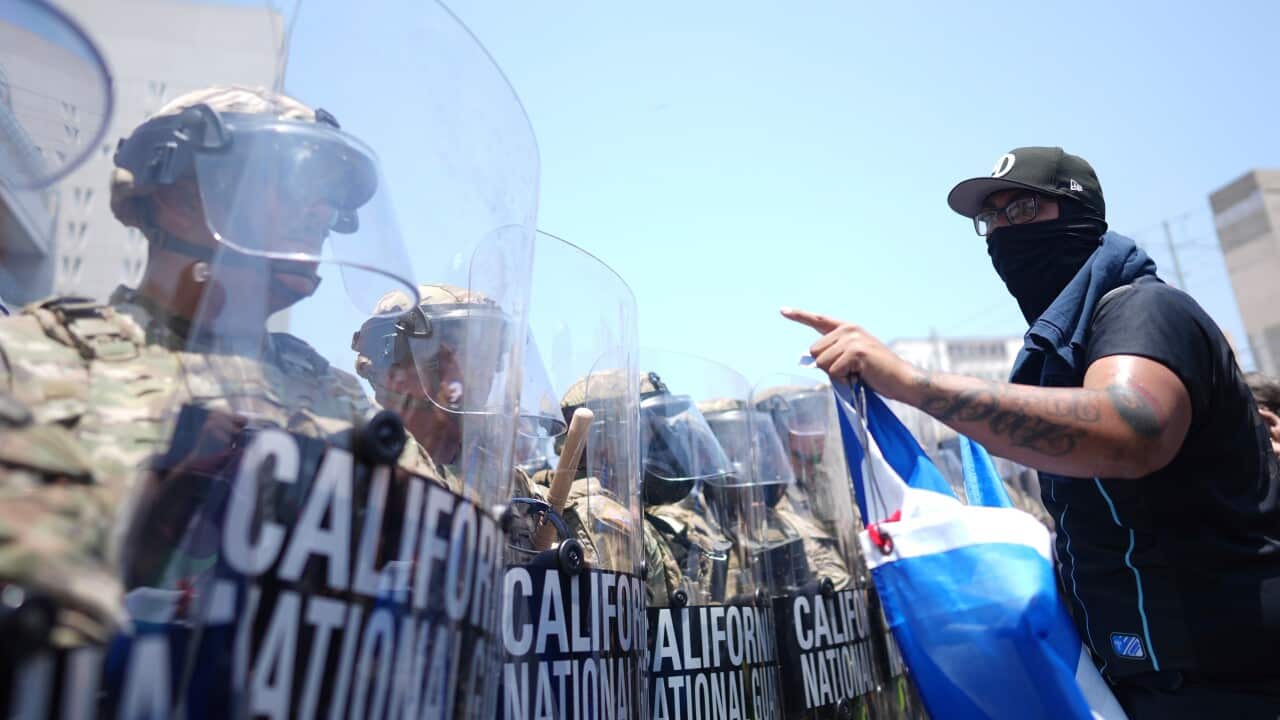 A man in a black t-shirt, cap, and face mask confronts a line of national guard troops holding riot shields.