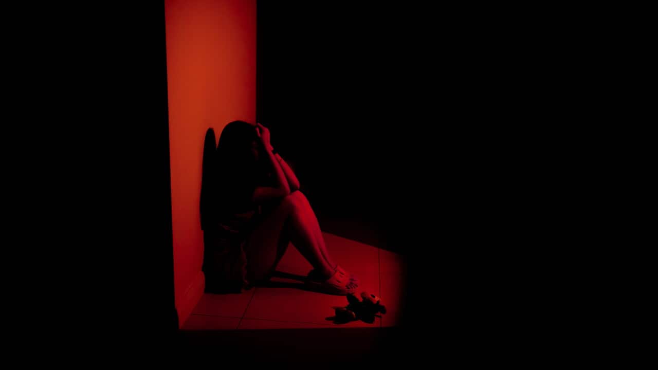 A woman sits on the floor, looking distressed, bathed in a red light in a dark room.