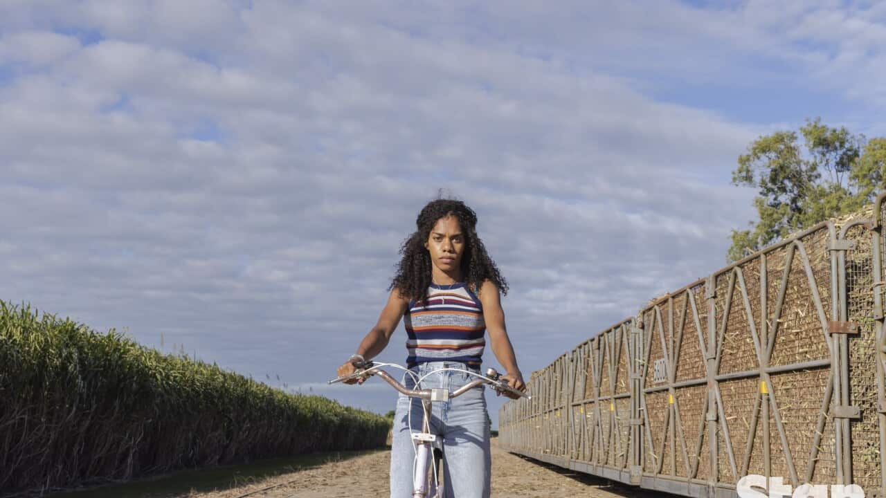 Talijah Blackman-Corowa in jeans and a striped singlet on a pushbike stops between a canefield and cane train