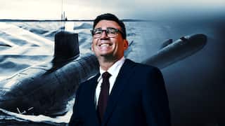 A man wearing a suit and glasses smiles. Behind him, there's the Union Jack flag and nuclear submarines moving through the ocean.