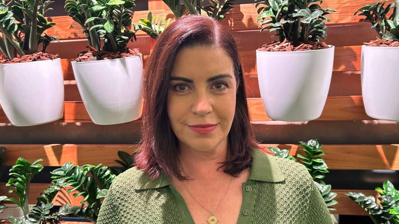 a woman with dark red hair and green top stands in front of a wall of plants