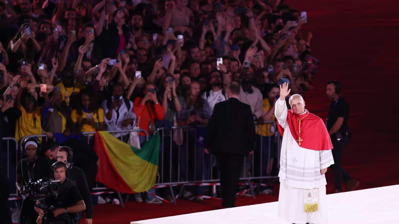 Pope Leo on a stage in front of a large group of people, holding up his left hand and waving.