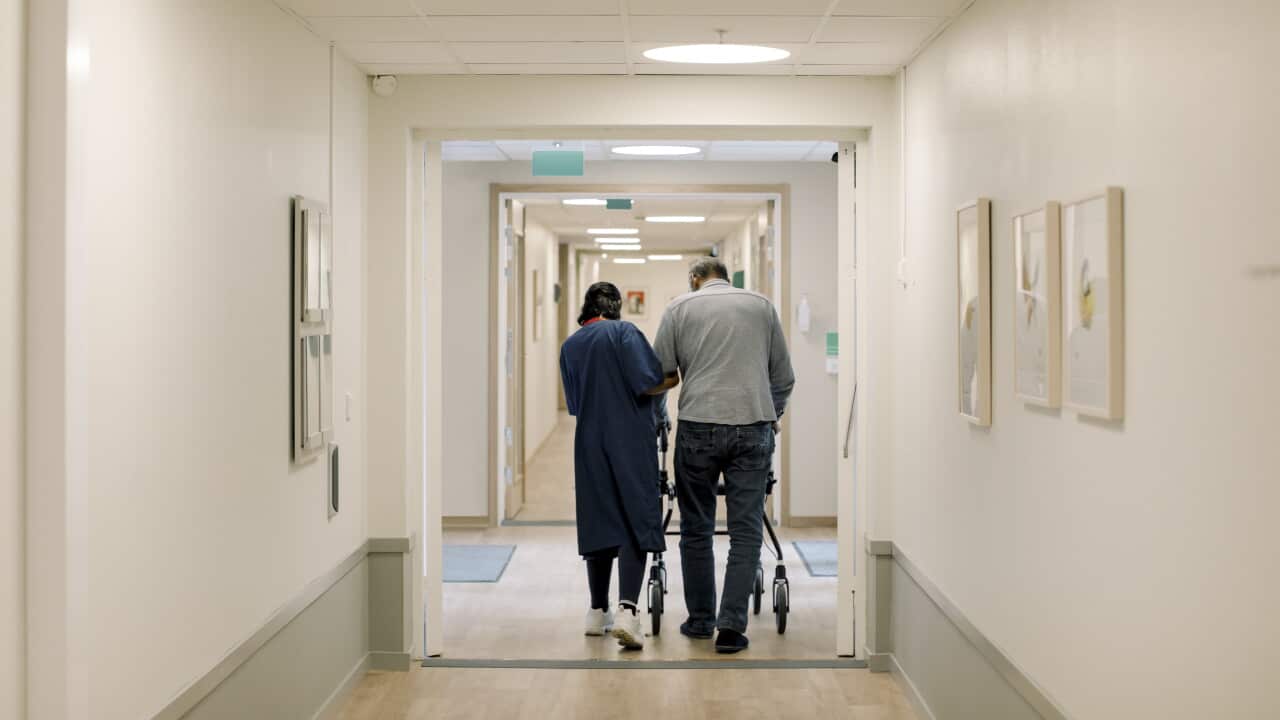 Two people walk down a healthcare facility corridor using wheeled walkers, with framed artwork on the walls and overhead lights lining the hallway.