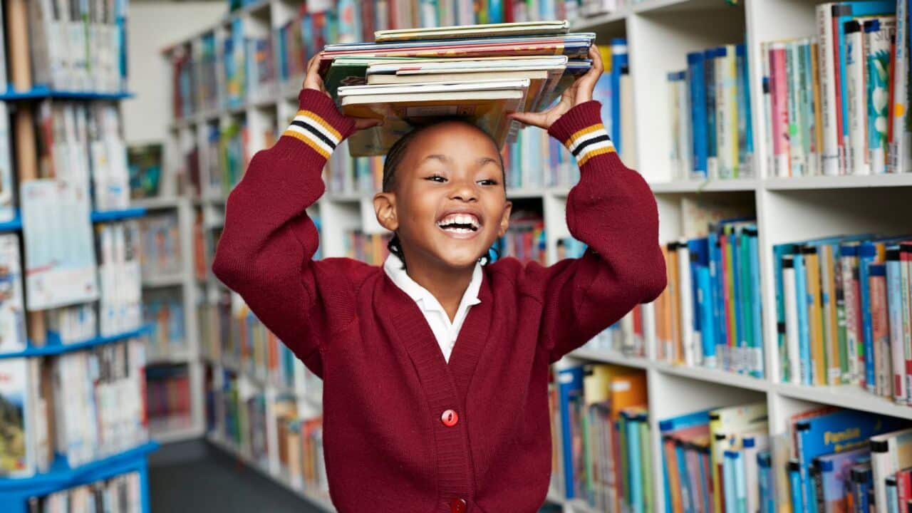 Cute schoolgirl smiling & balancing stack of books on the head at library