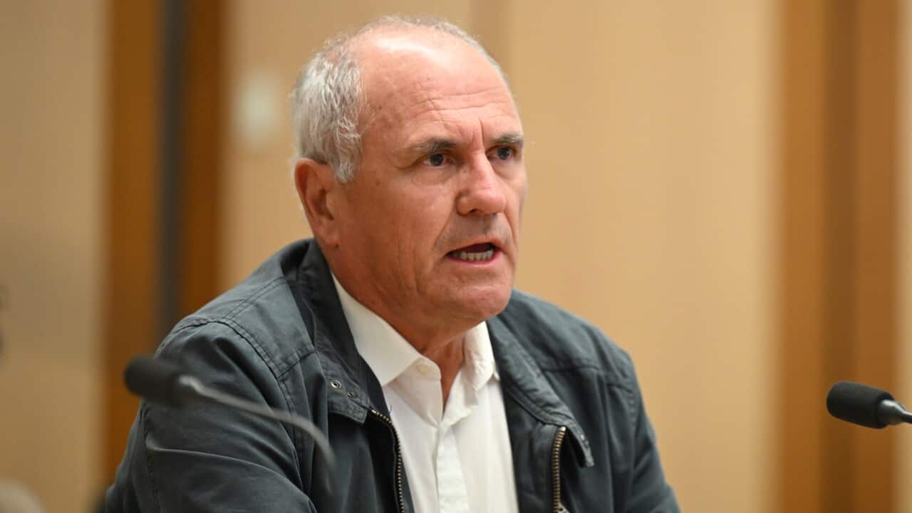 A man speaking while seated in front of a microphone. He looks frustrated and is wearing a faded black jacket and white collared shirt.
