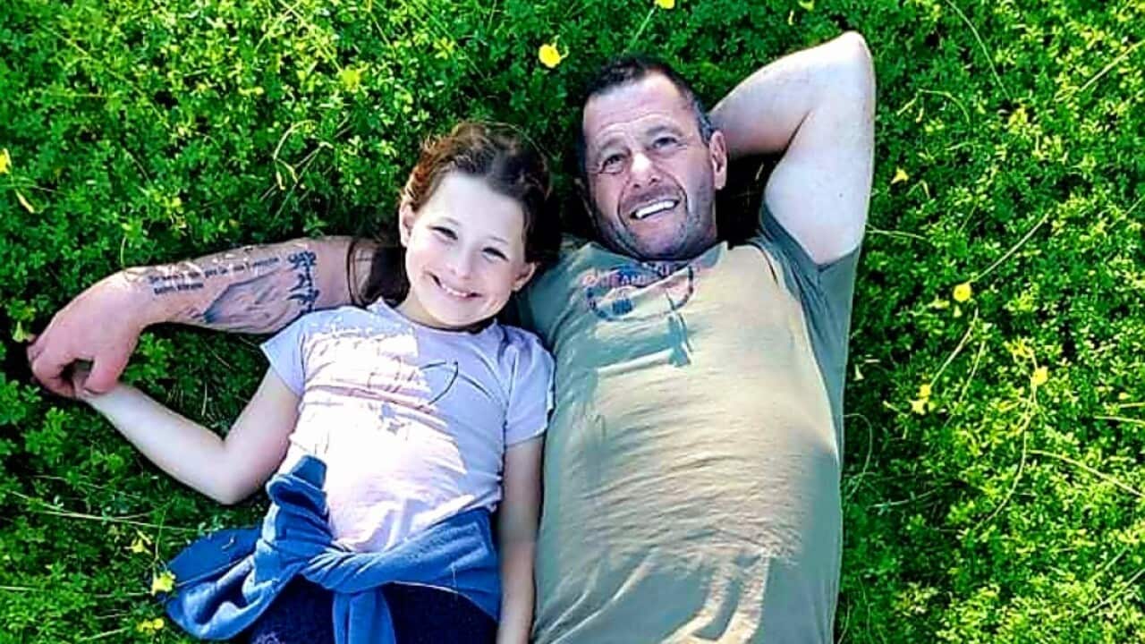 A man and his daughter lie on the grass together, his arm around her as they both smile.