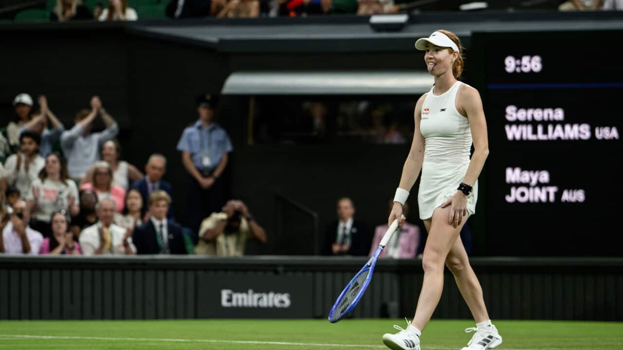 Maya Joint, a tennis player dressed in white and wearing sneakers, stands on a green tennis court with her tongue sticking out. A nearby crowd watches her play.
