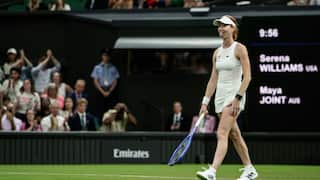 Maya Joint, a tennis player dressed in white and wearing sneakers, stands on a green tennis court with her tongue sticking out. A nearby crowd watches her play.
