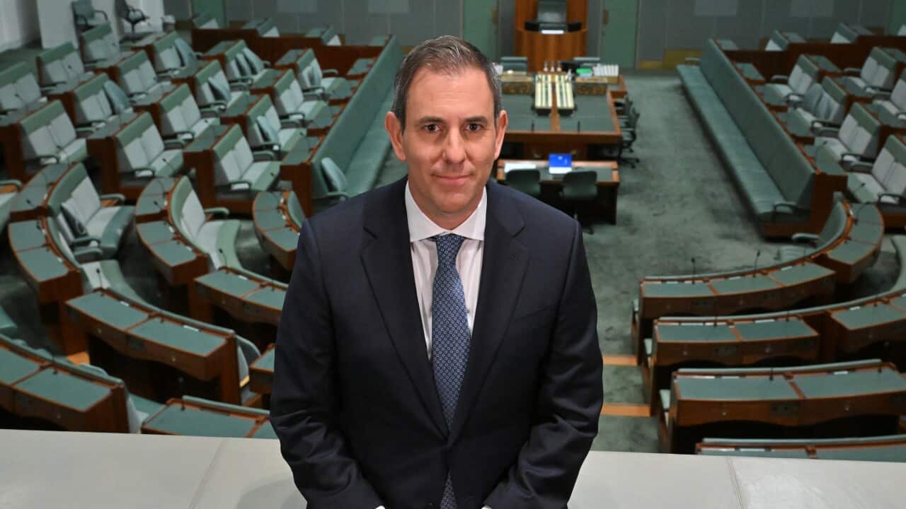 Jim Chalmers wearing a dark suit and tie. He is leaning on a barrier. Behind him is a chamber featuring green benches