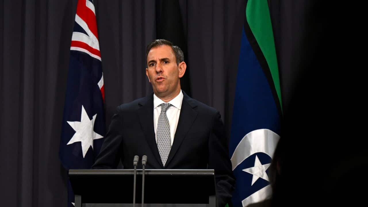 JIM CHALMERS speaking at a presser in front of a podium while wearing a suit, with the Australian and Torres Strait Islander flag behind him