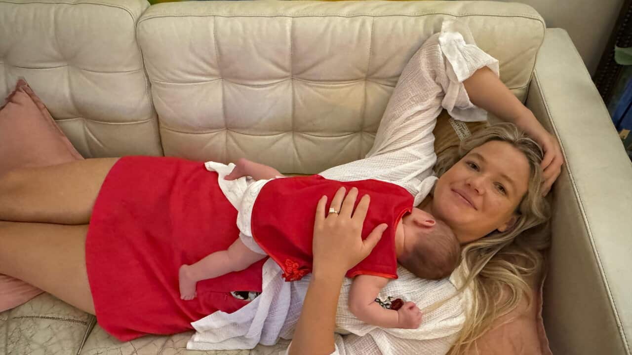 A young blonde white woman lying on a lounge and smiling. She is holding a baby in her arms.