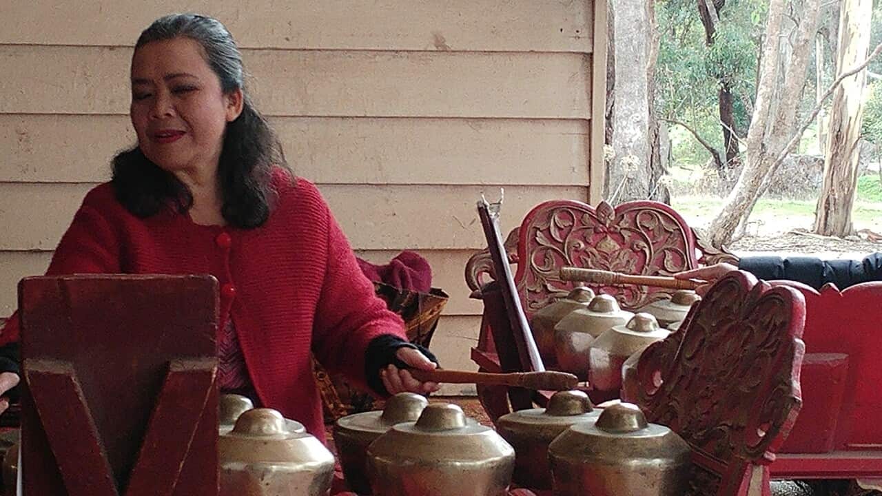 Nita plays the gamelan outside in front of weatherboard house with trees in background