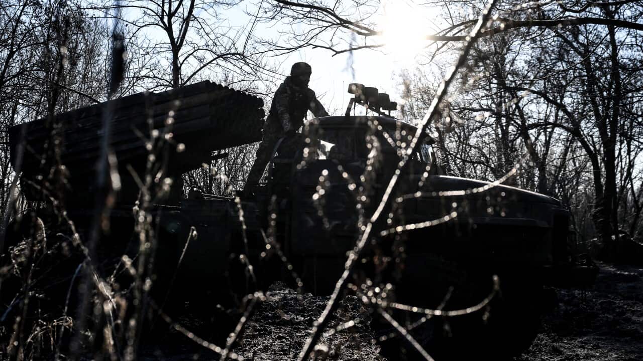 A soldier stands atop a truck-mounted multiple rocket launcher system positioned in a wooded area, silhouetted against a bright, low-hanging sun.