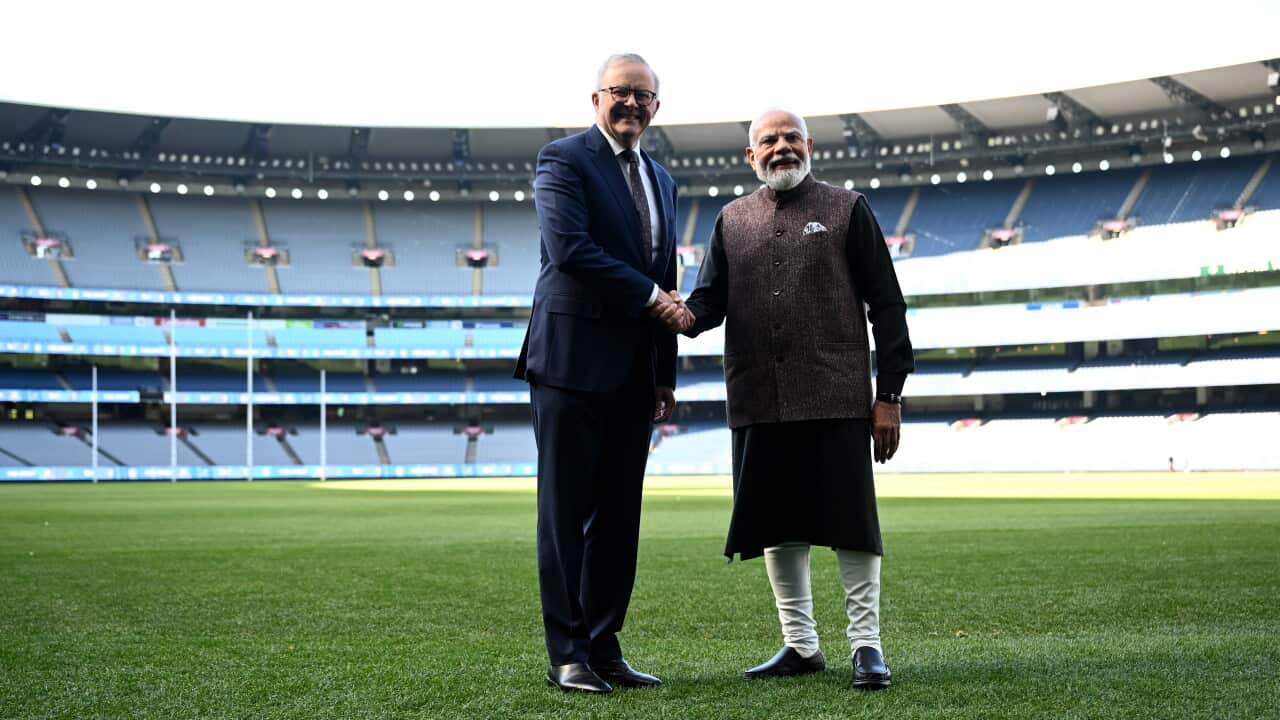 Australian Prime Minister Anthony Albanese and Indian Prime Minister Narendra Modi smile and shake hands while standing on a grassy sports field inside a large, empty stadium.