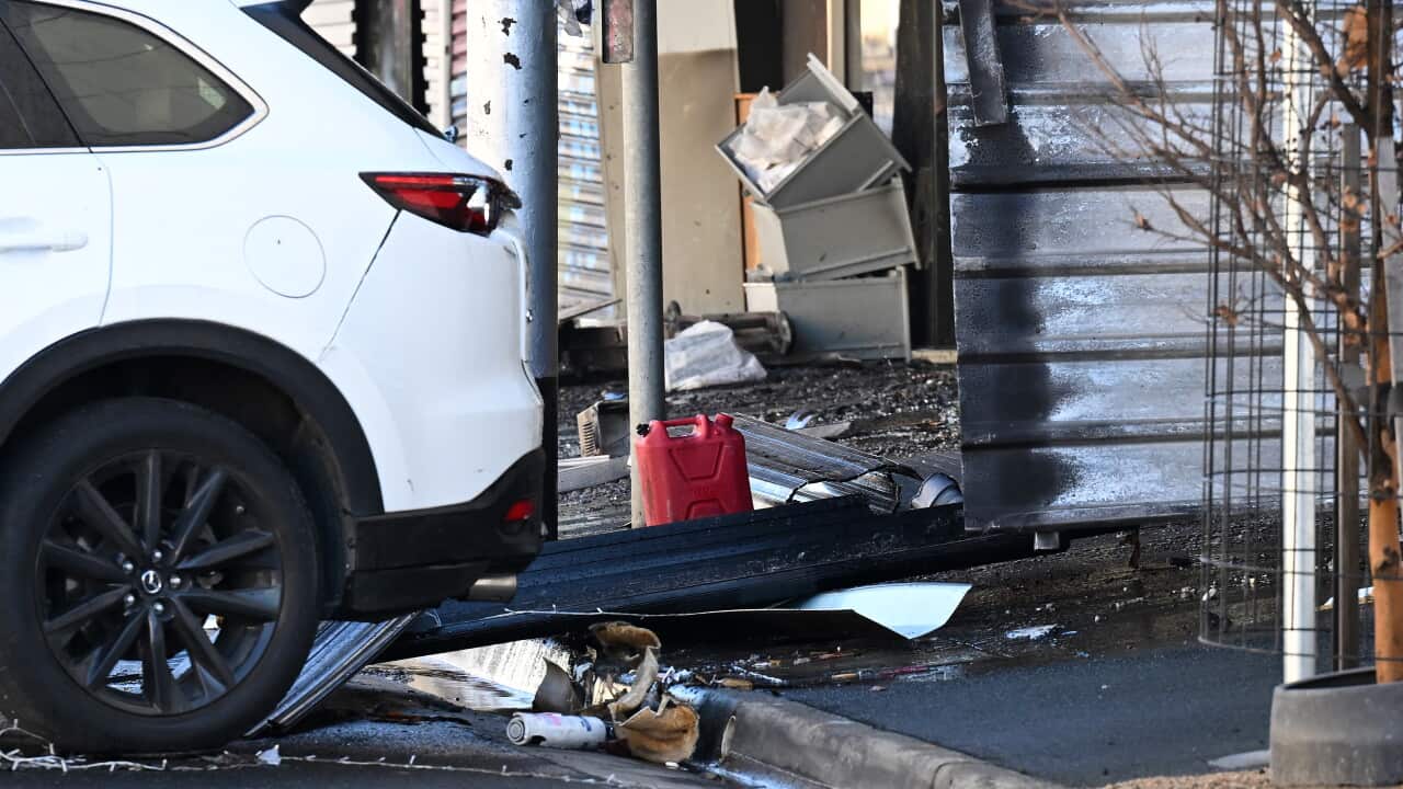A white SUV parked next to a storefront showing signs of fire damage, with debris, a red jerrycan, and charred building materials scattered on the ground.