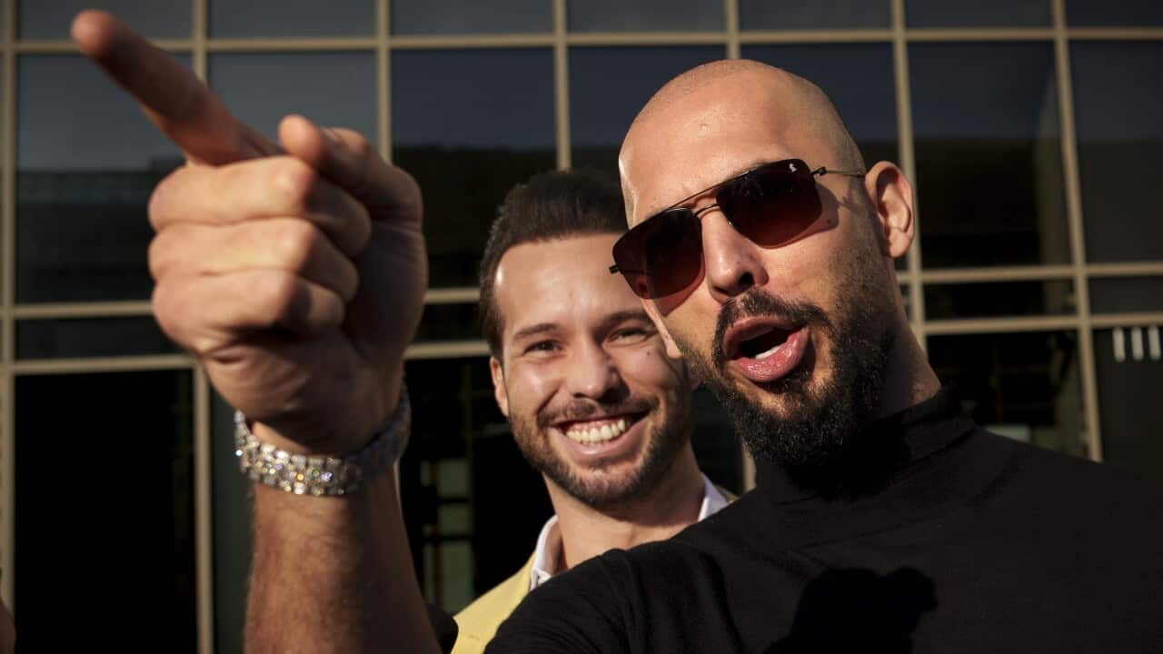 A man wearing sunnies is pointing his finger, with another man standing behind him and grinning.
