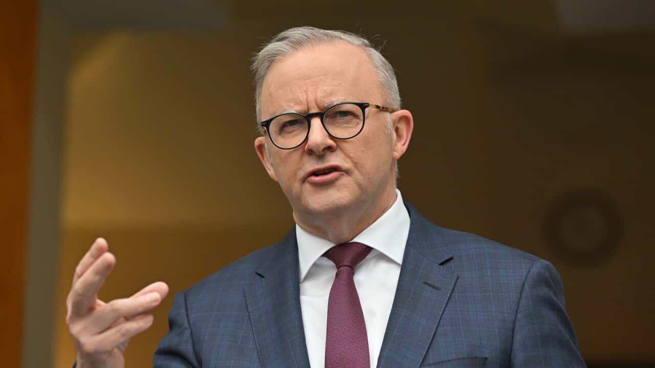 Anthony Albanese, wearing a dark blue blazer, white shirt, maroon tie, and glasses, is speaking with one hand raised.
