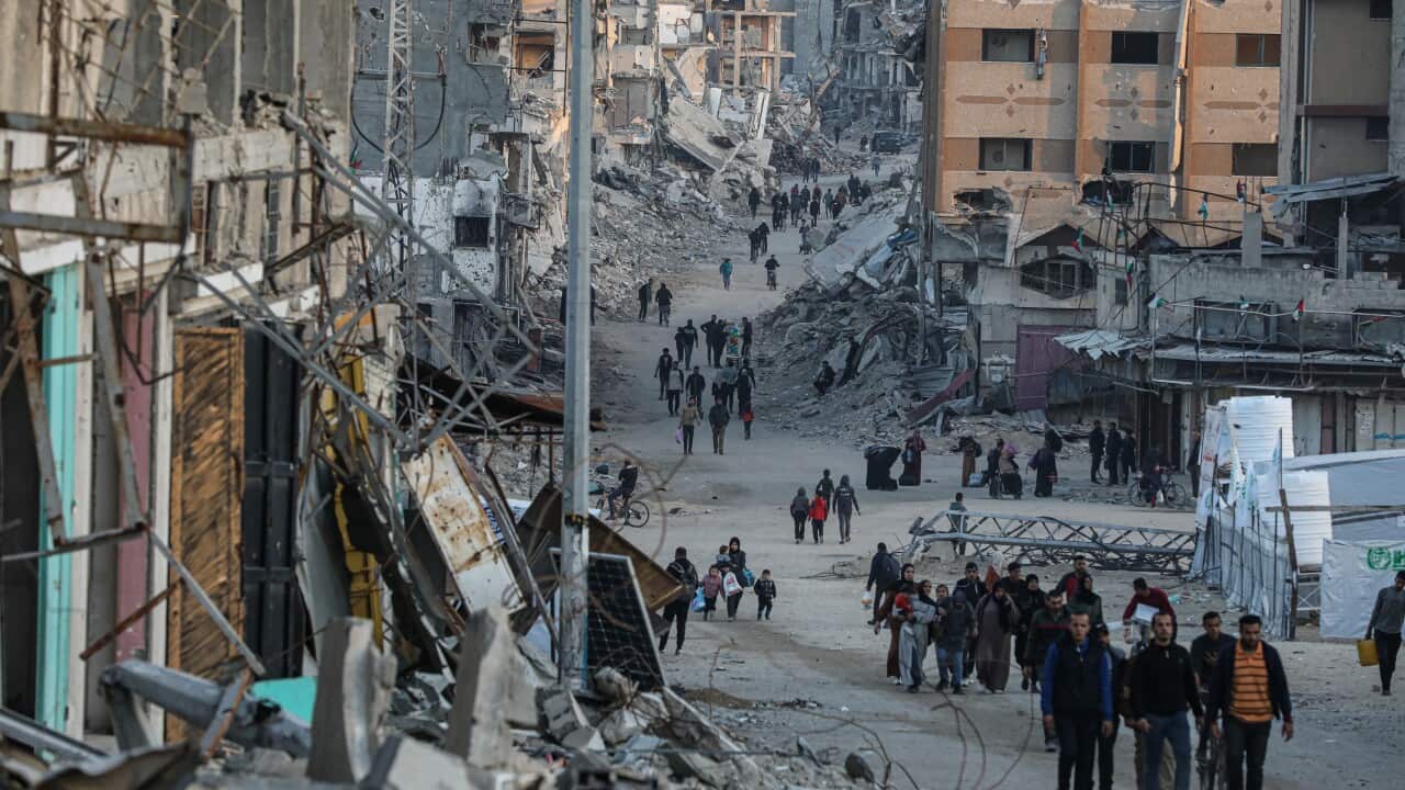 Dozens of people walk through a street lined with destroyed buildings carrying their belongings.