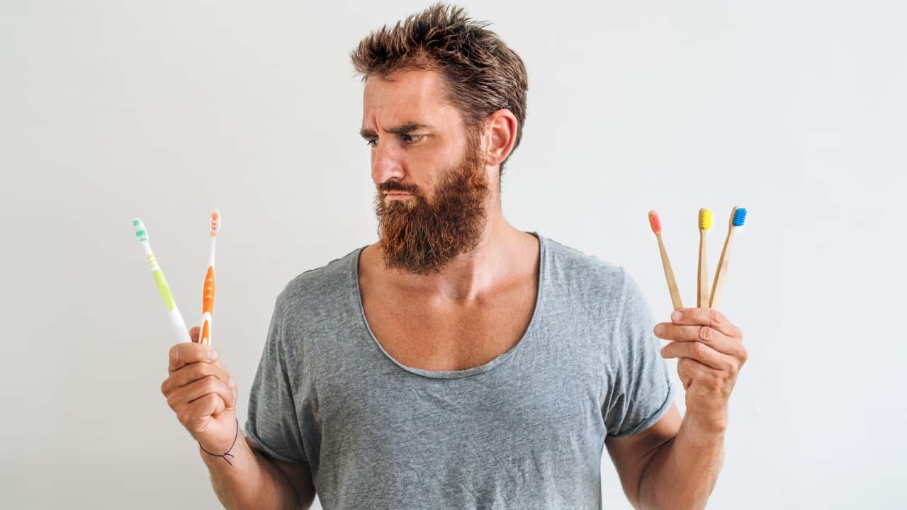 A front view portrait of man indoors holding plastic and ecological tooth brushes.