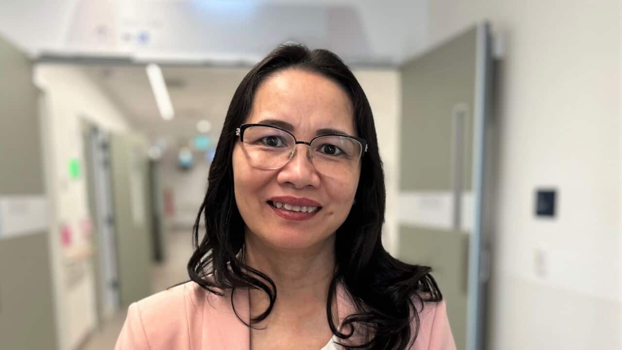 A woman in a pink jacket smiles to camera in a hospital corridor.