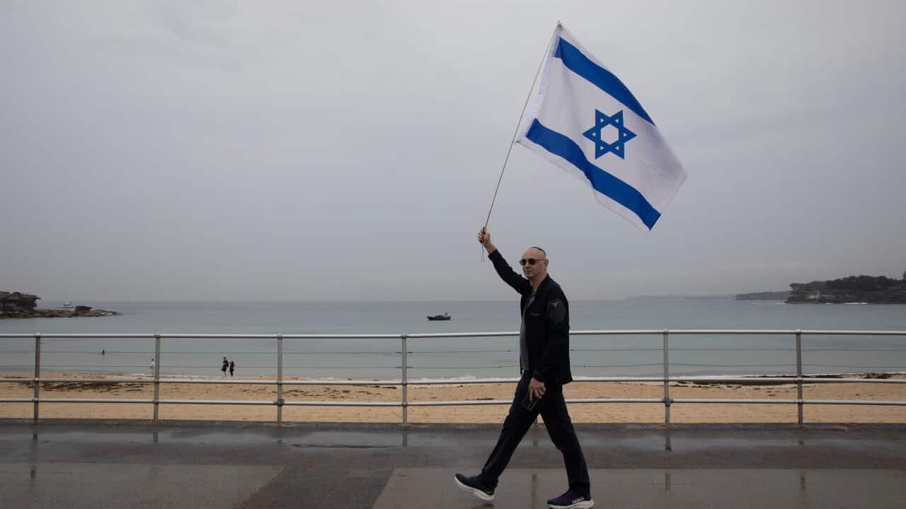 A man holding an Israeli flag as he walks along a promenade, a beach is in the background.