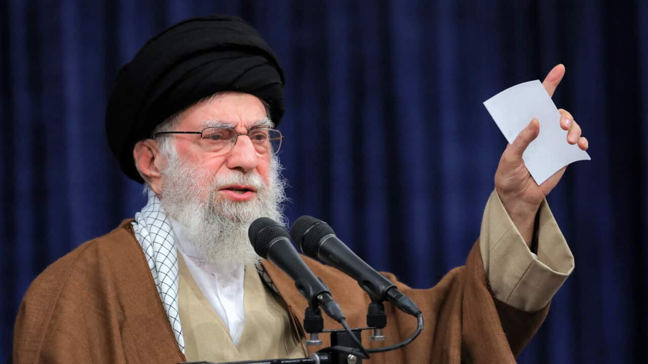 A man in an Islamic headdress raises his left index finger as he speaks in front of a microphone.