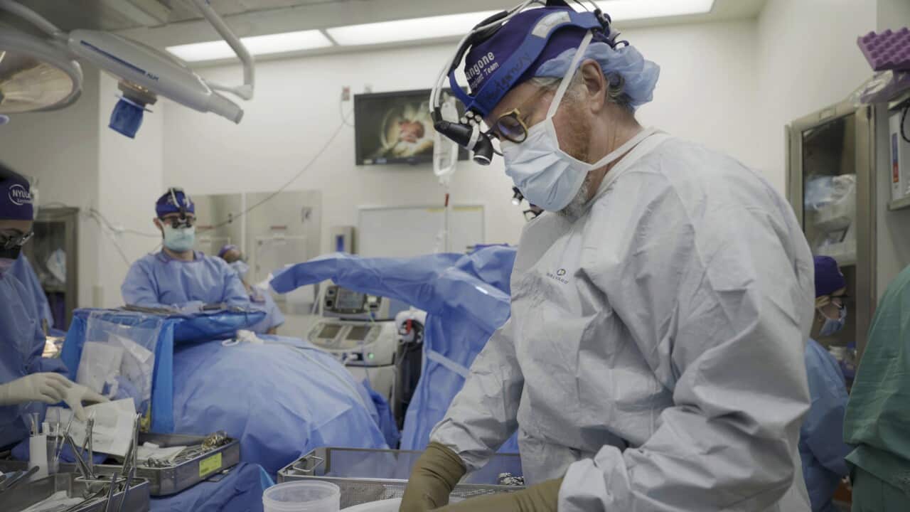 A person in medical wear moves something in a plastic container as others look on.