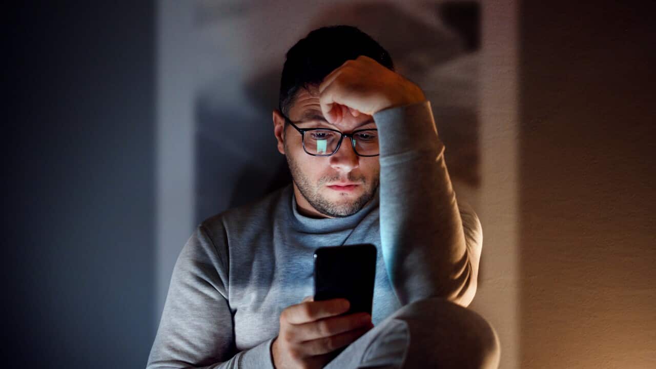A man sitting in a dark room with his hand on his head looking at a smartphone