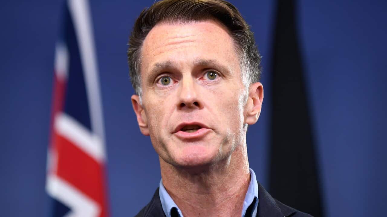 A man in a dark suit stands in front of a background with an Australian flag visible.
