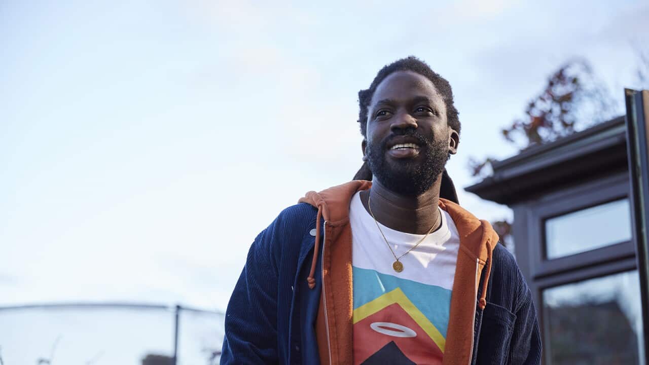 A man in a bright t-shirt and dark parka stands outdoors with a pale sky above, looking up and to the side.