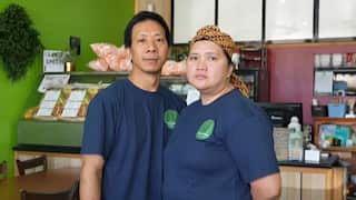 A couple wearing matching navy shirts stand side by side inside a small restaurant, with a food display and counter behind them.