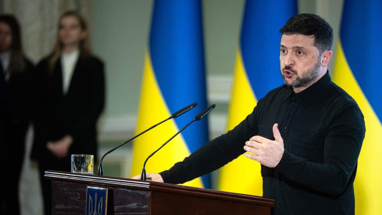 Ukrainian President Volodymyr Zelenskyy speaking before a lecturn. Ukrainian flags are hanging in the background.