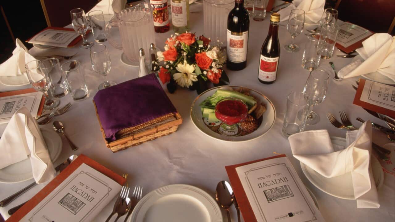 A table with covered in a white tablecloth and featuring settings of white plates with knives, spoons and forks, white serviettes on plates, some foot on a plate, flowers and bottles of wine.