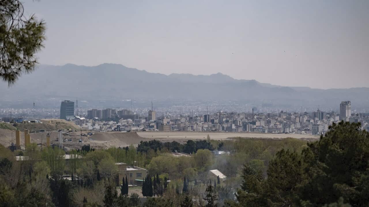 A high-angle cityscape of Tehran shows a dense sprawl of buildings and a few modern skyscrapers nestled between lush green trees in the foreground and the hazy, rugged Alborz Mountains in the distance.