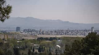 A high-angle cityscape of Tehran shows a dense sprawl of buildings and a few modern skyscrapers nestled between lush green trees in the foreground and the hazy, rugged Alborz Mountains in the distance.